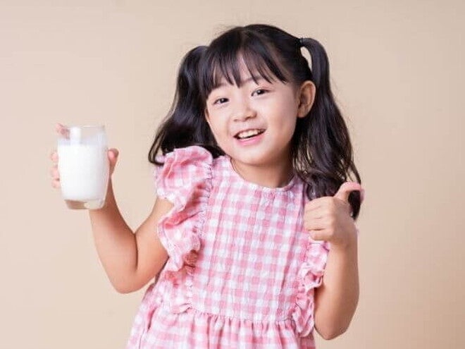 Menina sorrindo, vestindo um vestido xadrez rosa e branco, segurando um copo de leite e fazendo um joinha Menina sorrindo, vestindo um vestido xadrez rosa e branco, segurando um copo de leite e fazendo um joinha