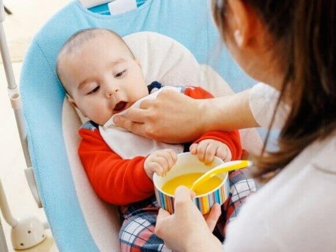 Bebê sentado em uma cadeirinha, com as mãos em um potinho de comida, sendo limpo por um adulto utilizando uma fralda de boca. Bebê sentado em uma cadeirinha, com as mãos em um potinho de comida, sendo limpo por um adulto utilizando uma fralda de boca.
