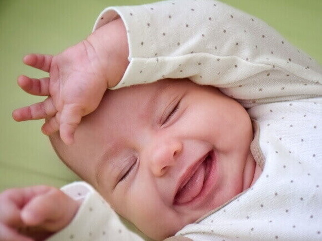 Bebê sorrindo de olhos fechados, com a mão perto do rosto, vestindo roupa branca com bolinhas cinzas. Bebê sorrindo de olhos fechados, com a mão perto do rosto, vestindo roupa branca com bolinhas cinzas.