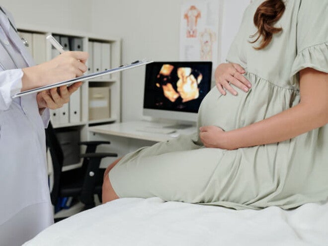 Gestante em consultório médico, sentada sob uma maca de atendimento, juntamente com um médico