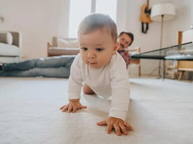 Engatinhar é bom? Incentive o bebê antes dos primeiros passos Bebê engatinhando no chão da sala de casa, com a supervisão do pai olhando do canto da sala