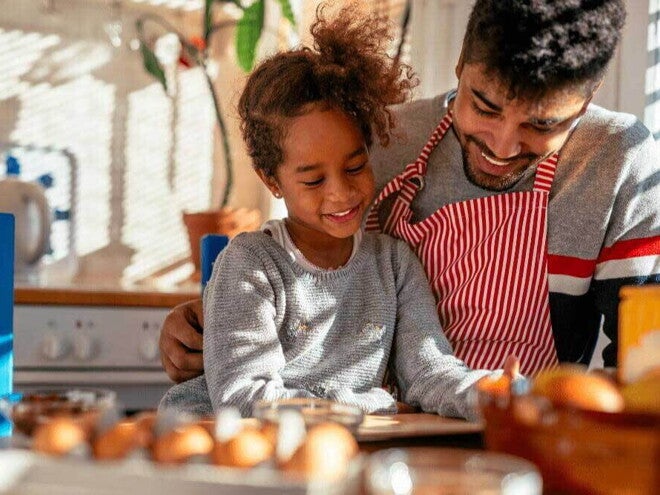 A relação de pai e filho é ilustrada por uma imagem onde a menina é abraçada pelo pai, que veste avental, enquanto eles olham para uma mesa de café da manhã com alguns alimentos. A relação de pai e filho é ilustrada por uma imagem onde a menina é abraçada pelo pai, que veste avental, enquanto eles olham para uma mesa de café da manhã com alguns alimentos.