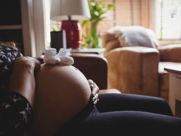 imagem com foco na barriga de uma mulher grávida, com dois sapatinhos de bebê na cor branca posicionados acima da barriga