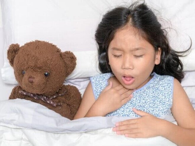 Uma menina deitada na cama com expressão de desconforto, segurando a garganta, ao lado de um ursinho de pelúcia. Uma menina deitada na cama com expressão de desconforto, segurando a garganta, ao lado de um ursinho de pelúcia.