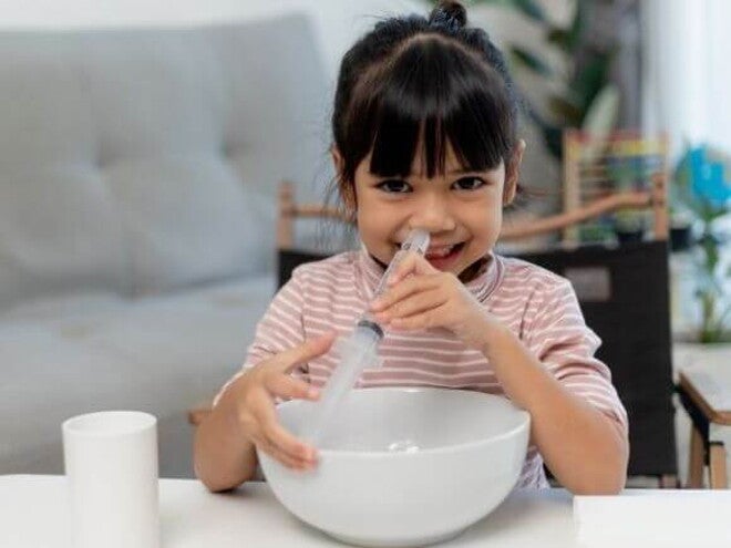 Uma menina sentada em uma mesa, com um pote em sua frente, segurando uma seringa no nariz