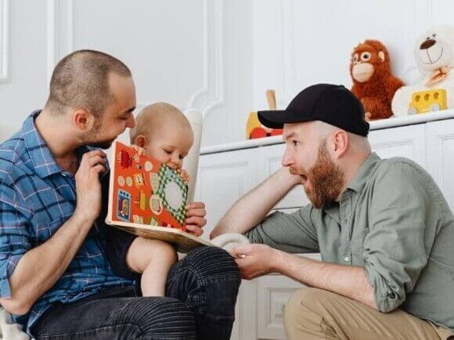 Um casal homoafetivo de dois pais, sorrindo e interagindo com um bebê, sentado no colo de um dos homens. Um casal homoafetivo de dois pais, sorrindo e interagindo com um bebê, sentado no colo de um dos homens.