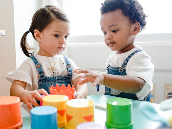 imagem de duas crianças pequenas vestindo macacão jeans e camiseta branca, brincando com brinquedos pedagógicos.