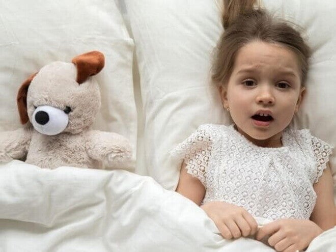 Uma menina assustada está deitada na cama, coberta por um lençol, com expressão de medo, ao lado de um cachorro de pelúcia. Uma menina assustada está deitada na cama, coberta por um lençol, com expressão de medo, ao lado de um cachorro de pelúcia.