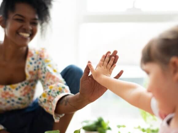 mulher negra com um largo sorriso e uma blusa floral dá um "toca aqui" a uma criança pequena, que estende a mão em resposta