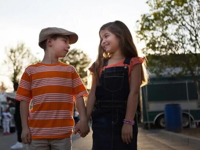 Imagem de um menino e uma menina de mãos dadas se olhando. O menino usa uma boina e uma camisa listrada laranja e branco e a menina está de cabelo solto, usando um macacão escuro e sorrindo Imagem de um menino e uma menina de mãos dadas se olhando. O menino usa uma boina e uma camisa listrada laranja e branco e a menina está de cabelo solto, usando um macacão escuro e sorrindo