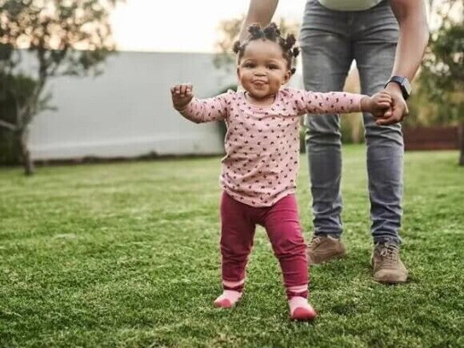 O que é coordenação motora e quais suas fases nas crianças Menina aprendendo a andar e ganhando coordenação motora. Ela caminha no quintal orientada pela mãe, que segura a sua mão esquerda.