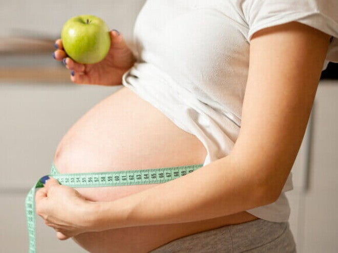 Mulher grávida segurando uma maçã em uma mão e uma fita métrica para costura na outra, ao redor da barriga. Mulher grávida segurando uma maçã em uma mão e uma fita métrica para costura na outra, ao redor da barriga.