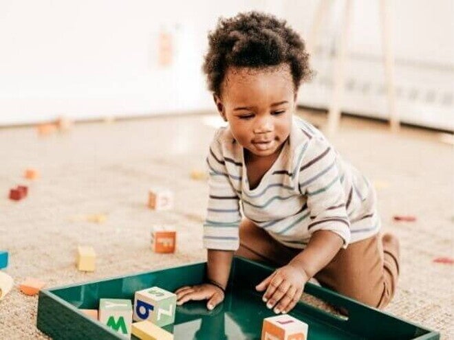 Criança pequena brincando no chão com blocos de montar coloridos em uma bandeja verde, com vários brinquedos espalhados ao redor sobre um tapete bege em um ambiente iluminado. Criança pequena brincando no chão com blocos de montar coloridos em uma bandeja verde, com vários brinquedos espalhados ao redor sobre um tapete bege em um ambiente iluminado.