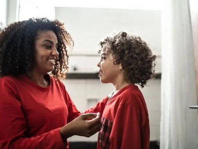 Palavras mágicas Mulher sorrindo com seu filho, ambos usando um pijama vermelho e conversando. Ao fundo, um quarto.