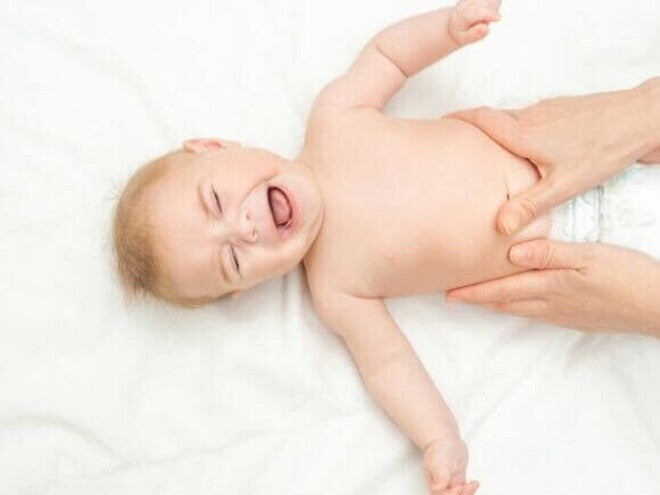Bebê deitado de costas em uma superfície branca, chorando, enquanto um adulto realiza uma massagem suave em sua barriga. Bebê deitado de costas em uma superfície branca, chorando, enquanto um adulto realiza uma massagem suave em sua barriga.