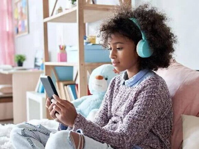 Pré-adolescência #ParaTodosVerem Menina negra pré-adolescente, com cabelo cacheado, solto, usando fones de ouvido azuis enquanto olha a tela do celular. Ela está sentada em uma cama com almofadas e veste blusão com calça jeans. O fundo da foto é um quarto de menina.