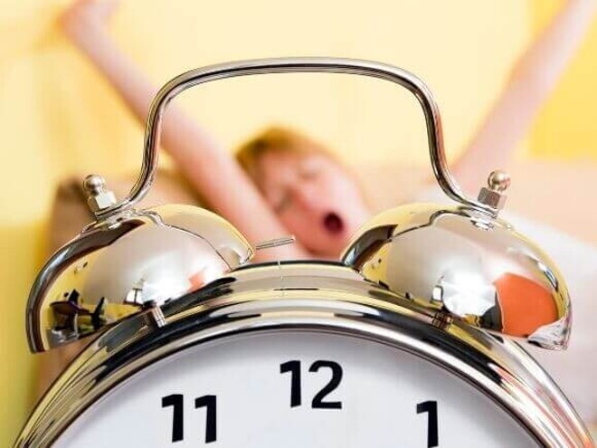 Relógio despertador prateado em primeiro plano, com uma criança ao fundo bocejando e se espreguiçando em uma cama Relógio despertador prateado em primeiro plano, com uma criança ao fundo bocejando e se espreguiçando em uma cama