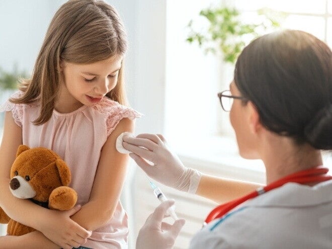 Uma menina segura um ursinho de pelúcia enquanto uma profissional de saúde, prepara o braço da menina para uma injeção com um algodão. Uma menina segura um ursinho de pelúcia enquanto uma profissional de saúde, prepara o braço da menina para uma injeção com um algodão.