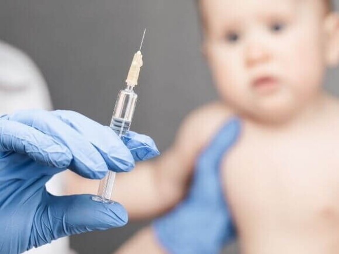 Mão com luva cirúrgica segurando seringa com a vacina da coqueluche, enquanto bebê aguarda a aplicação Mão com luva cirúrgica segurando seringa com a vacina da coqueluche, enquanto bebê aguarda a aplicação