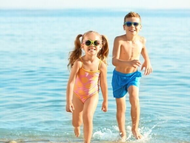 Duas crianças sorrindo e correndo na praia, usando óculos de sol e roupa de banho. Duas crianças sorrindo e correndo na praia, usando óculos de sol e roupa de banho.