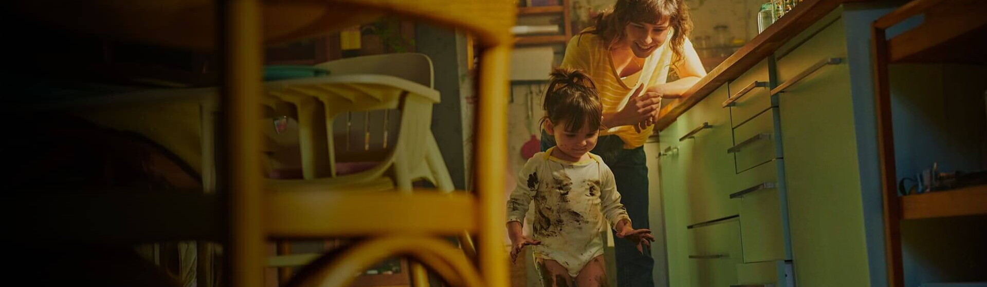 Mãe vestida com uma blusa amarela, encostada na bancada da cozinha, observa, sorridente, a filha que está suja com tinta nas mãos, nas pernas e na roupa Mãe vestida com uma blusa amarela, encostada na bancada da cozinha, observa, sorridente, a filha que está suja com tinta nas mãos, nas pernas e na roupa