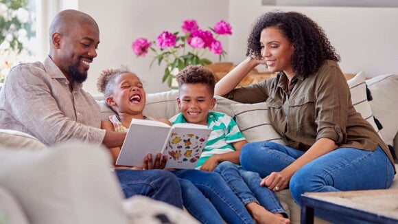 Uma família, formada por pai, mãe e dois filhos, sentada no sofá, lendo um livro juntos e sorrindo. Uma família, formada por pai, mãe e dois filhos, sentada no sofá, lendo um livro juntos e sorrindo.