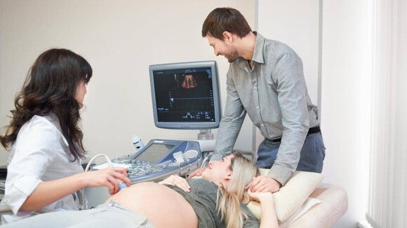 Gestante deitada em uma maca realizando exame pré-natal com ultrassom, acompanhada por um profissional de saúde e uma pessoa ao seu lado oferecendo apoio. Gestante deitada em uma maca realizando exame pré-natal com ultrassom, acompanhada por um profissional de saúde e uma pessoa ao seu lado oferecendo apoio.