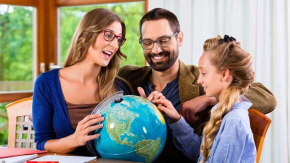 Um homem e uma mulher sorridentes, olham para uma menina que aponta para um globo terrestre sobre uma mesa. Um homem e uma mulher sorridentes, olham para uma menina que aponta para um globo terrestre sobre uma mesa.