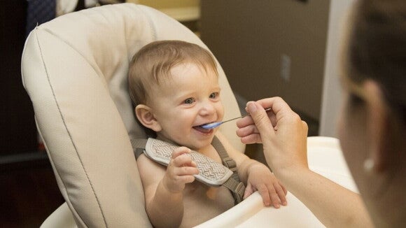 Bebê sorridente sentado em uma cadeira de alimentação, sendo alimentado por um adulto com uma colher de comida. Bebê sorridente sentado em uma cadeira de alimentação, sendo alimentado por um adulto com uma colher de comida.