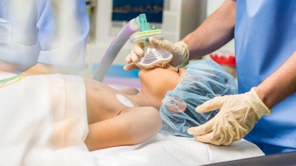 Uma criança deitada em uma mesa de cirurgia, com uma touca cirúrgica e uma máscara de anestesia sobre o rosto, segurada por uma pessoa usando luvas cirúrgicas. Uma criança deitada em uma mesa de cirurgia, com uma touca cirúrgica e uma máscara de anestesia sobre o rosto, segurada por uma pessoa usando luvas cirúrgicas.