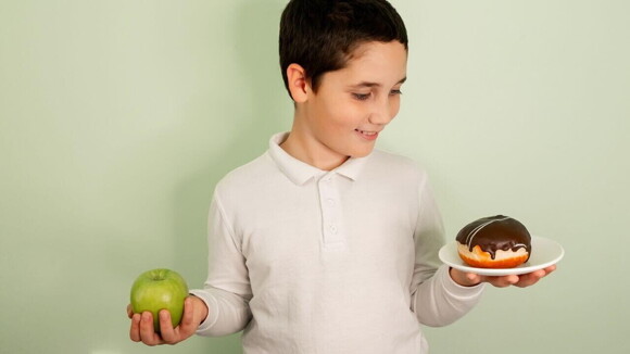 Menino escolhendo entre uma maçã e uma rosquinha, representando as escolhas alimentares e o colesterol infantil Menino escolhendo entre uma maçã e uma rosquinha, representando as escolhas alimentares e o colesterol infantil
