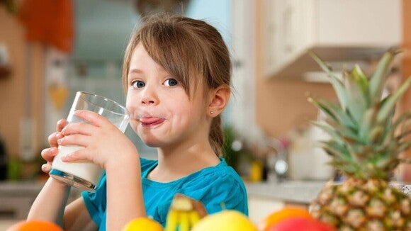 Menina bebendo um copo de leite, com frutas ao redor e uma cozinha ao fundo. Menina bebendo um copo de leite, com frutas ao redor e uma cozinha ao fundo.