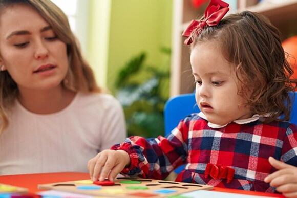 Criança com síndrome de down brincando com uma prancha de encaixe de formas ao lado de uma mulher