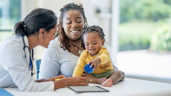 Médica conversando com mãe e bebê durante uma consulta pediátrica, com o bebê brincando com um cubo azul sobre a mesa. Médica conversando com mãe e bebê durante uma consulta pediátrica, com o bebê brincando com um cubo azul sobre a mesa.