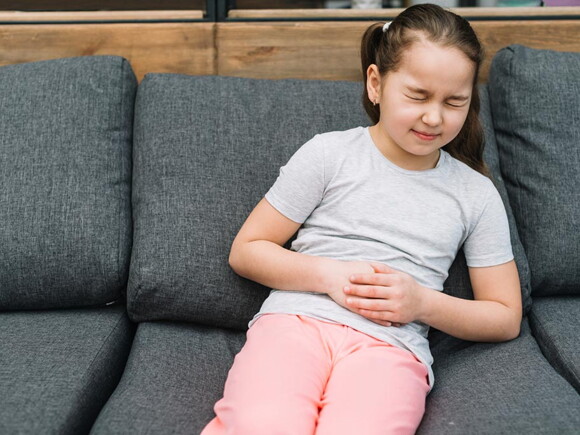 menina sentada ao sofá agarra a barriga com feição de dor