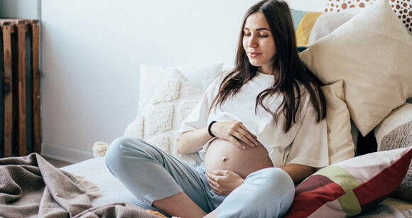 Gestante sentada na cama com as mãos sobre a barriga