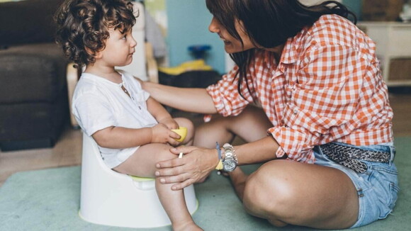 Bebê de cabelos cacheados sentado em um penico branco, e sua mãe sentada em sua frente usando camisa xadrez e short jeans. Bebê de cabelos cacheados sentado em um penico branco, e sua mãe sentada em sua frente usando camisa xadrez e short jeans.
