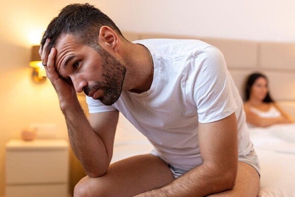 Imagem de um homem sentado na cama com a mão sobre a cabeça com semblante preocupado e esposa com os braços cruzados