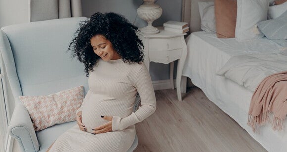 Imagem de uma gestante sentada em uma poltrona com as mãos sobre a barriga olhando para baixo sorrindo Imagem de uma gestante sentada em uma poltrona com as mãos sobre a barriga olhando para baixo sorrindo