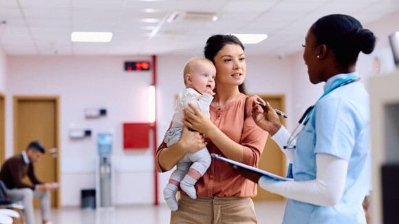 Uma mulher segurando um bebê nos braços em um hospital, conversando com uma profissional de saúde. Uma mulher segurando um bebê nos braços em um hospital, conversando com uma profissional de saúde.