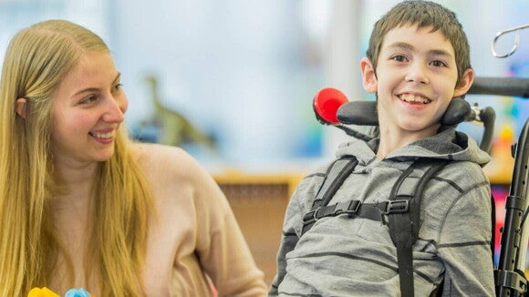 Um jovem com deficiência sorri enquanto está sentado em uma cadeira de rodas adaptada. Ao lado dele, uma mulher sorri olhando para ele. Um jovem com deficiência sorri enquanto está sentado em uma cadeira de rodas adaptada. Ao lado dele, uma mulher sorri olhando para ele.
