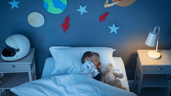 Um menino está dormindo em uma cama, abraçado a um ursinho de pelúcia, em um quarto com decoração temática de espaço. Um menino está dormindo em uma cama, abraçado a um ursinho de pelúcia, em um quarto com decoração temática de espaço.