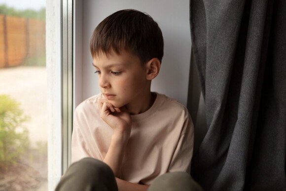 menino sentado junto a uma janela olha pensativamente para o lado de fora com seu queixo apoiado na mão