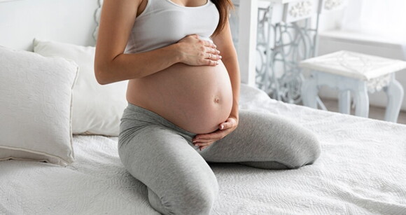 Mulher grávida sentada na cama com as mãos na barriga