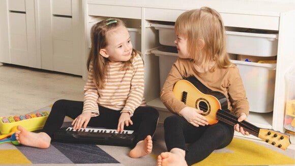Duas crianças sentadas em um tapete no chão. A criança da esquerda está com as mãos sobre um teclado de brinquedo, enquanto a criança da direita está segurando um violão de brinquedo. Duas crianças sentadas em um tapete no chão. A criança da esquerda está com as mãos sobre um teclado de brinquedo, enquanto a criança da direita está segurando um violão de brinquedo.