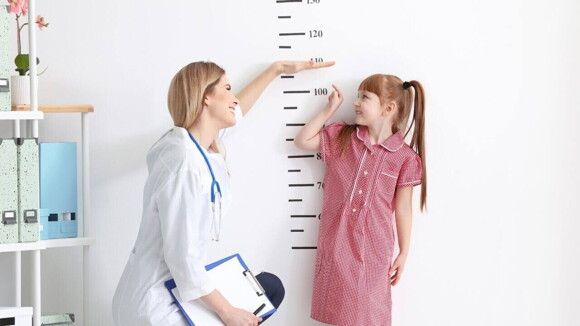 Uma profissional de saúde e uma menina, apontando para uma marcação de altura em um consultório médico. Uma profissional de saúde e uma menina, apontando para uma marcação de altura em um consultório médico.