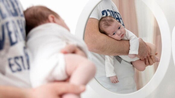 Bebê pequeno deitado de bruços no braço de um adulto, refletido em um espelho redondo. Bebê pequeno deitado de bruços no braço de um adulto, refletido em um espelho redondo.