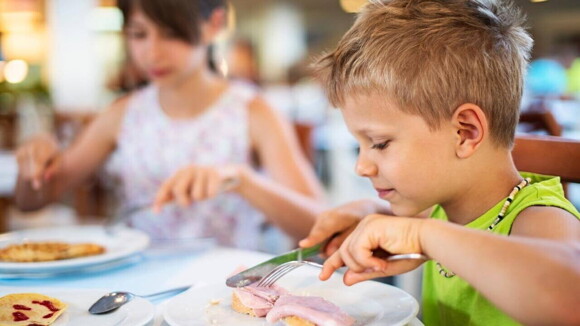 Criança comendo em uma mesa de refeição, concentrada enquanto corta uma fatia de pão com presunto. Ao fundo, outra criança desfocada também faz sua refeição Criança comendo em uma mesa de refeição, concentrada enquanto corta uma fatia de pão com presunto. Ao fundo, outra criança desfocada também faz sua refeição