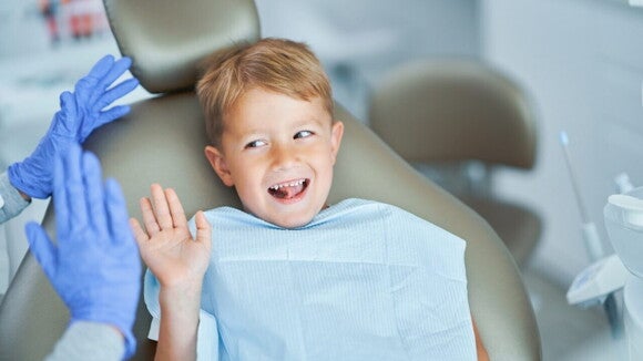 Um menino sorri e acena para alguém com luvas azuis, enquanto está sentado em uma cadeira de dentista. Um menino sorri e acena para alguém com luvas azuis, enquanto está sentado em uma cadeira de dentista.