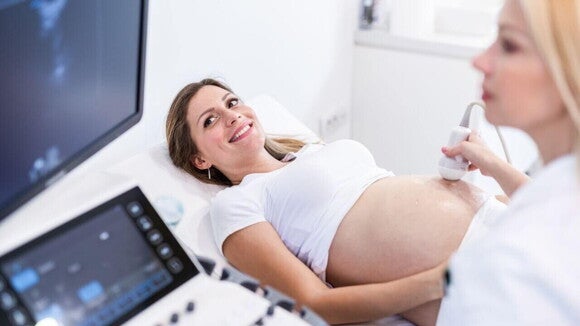 Mulher grávida deitada em uma maca realizando um exame de ultrassom abdominal, com profissional de saúde utilizando o transdutor sobre a barriga e aparelhos de ultrassom visíveis ao lado. Mulher grávida deitada em uma maca realizando um exame de ultrassom abdominal, com profissional de saúde utilizando o transdutor sobre a barriga e aparelhos de ultrassom visíveis ao lado.